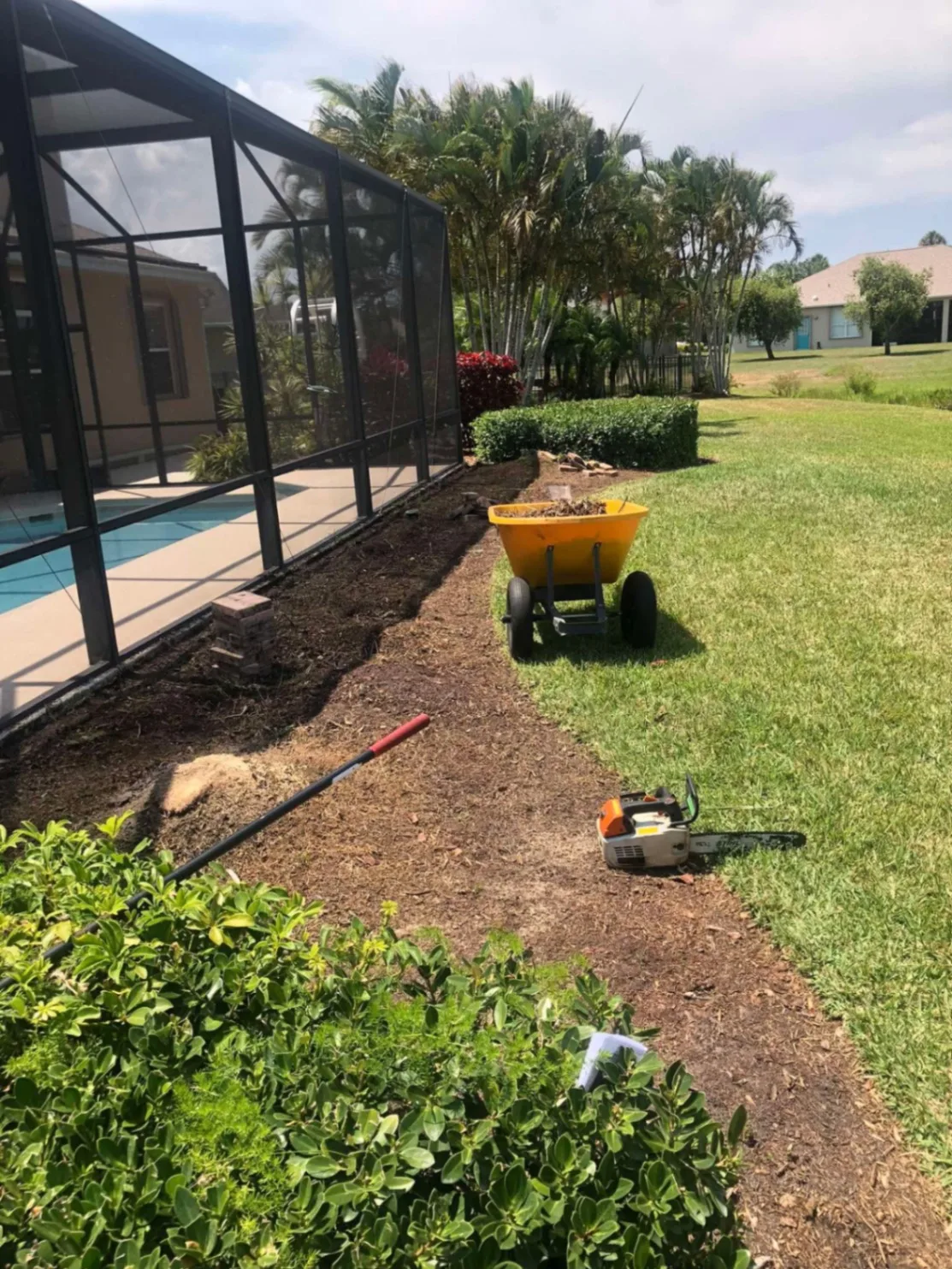 New bed being dug out along a pool enclosure with a wheelbarrow and tools